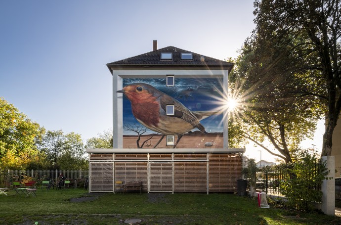 Das Wandbild von Denis Klatt zeigt ein überdimensionales Rotkehlchen, das in einer surrealen Szenerie aus Wüste und Meer eingebettet ist. (vergrößerte Bildansicht wird geöffnet)